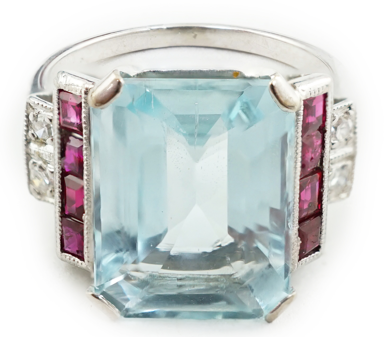 An Art Deco style platinum? and single stone emerald cut aquamarine set dress ring, with millegrain set ruby and diamond cluster stepped shoulders